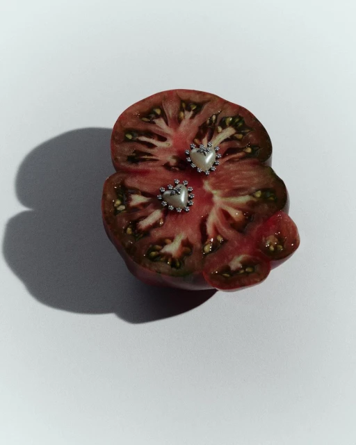 Artistic close-up of sliced tomato topped with two pearl and crystal heart earrings on a white background, casting a soft shadow.