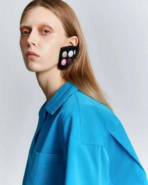 Blonde woman in a bright blue shirt, wearing an eye shadow palette as an earring, posing in front of a neutral-toned background.