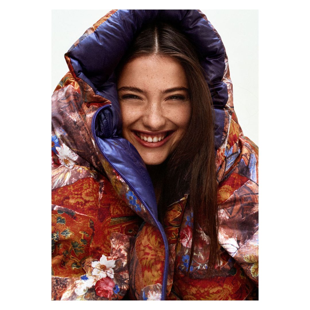 Smiling woman wearing a floral puffer jacket with a hood. Head and shoulders shot, indoors. Beauty and fashion portrait.