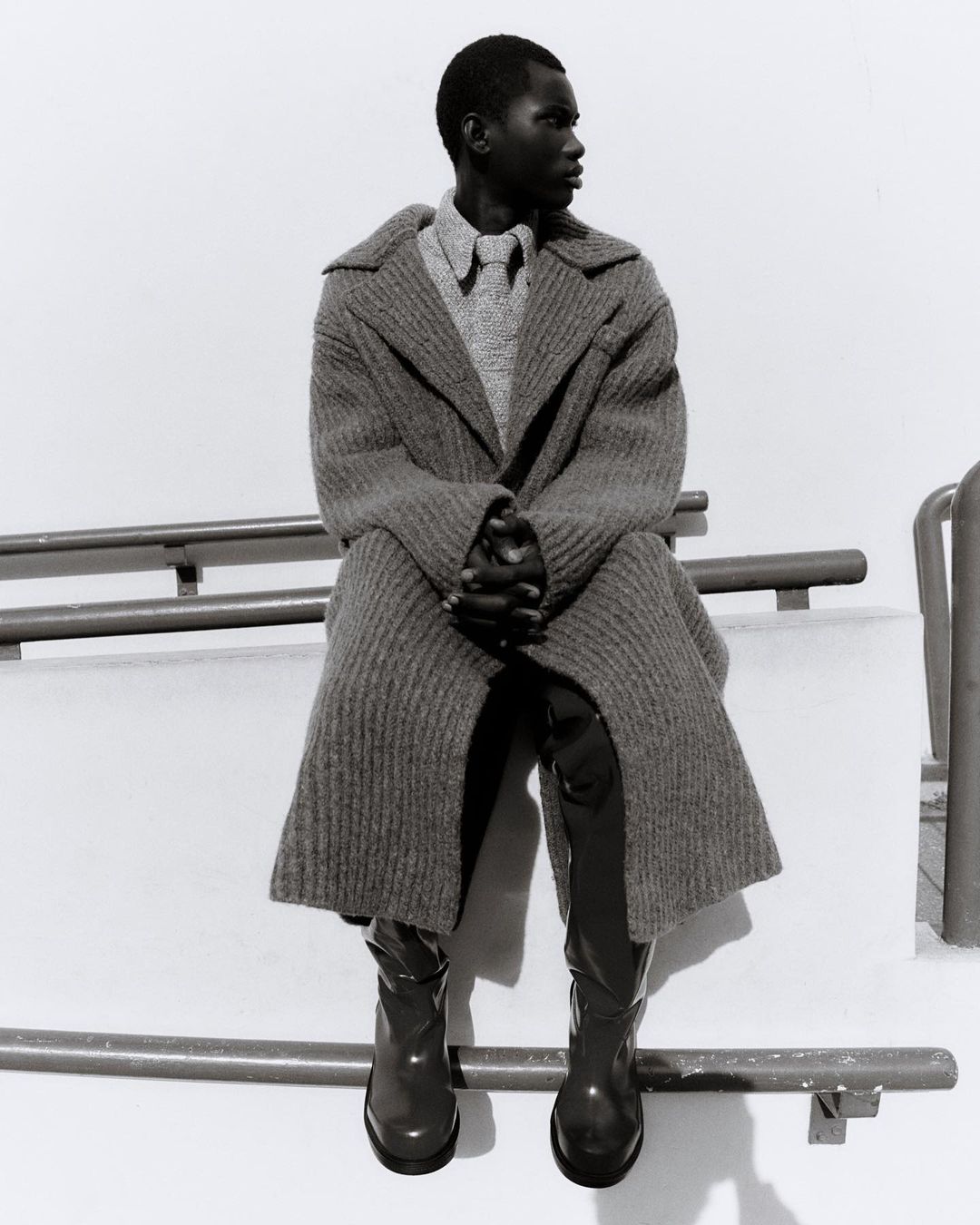 Black male fashion model in coat and boots sits on a railing in black and white. The model looks to the side.