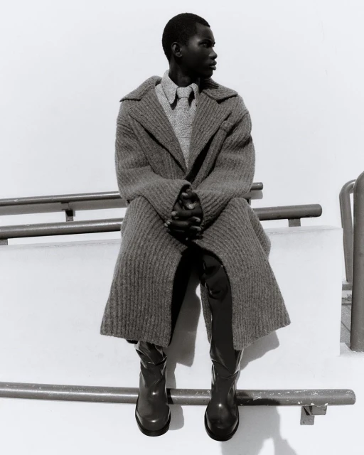 Black male fashion model in coat and boots sits on a railing in black and white. The model looks to the side.