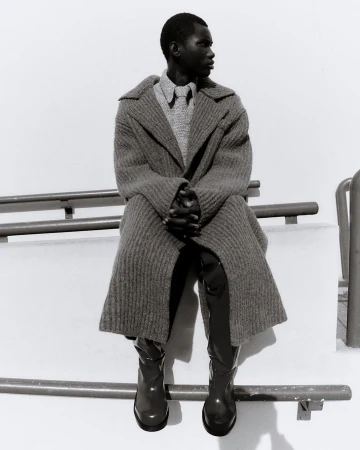 Black male fashion model in coat and boots sits on a railing in black and white. The model looks to the side.