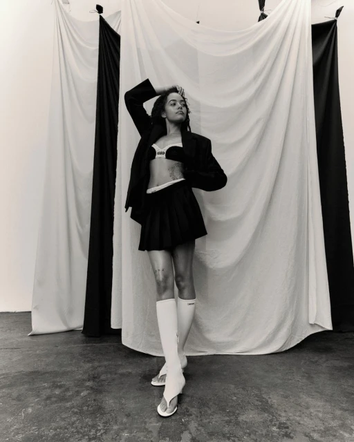 Monochrome portrait of a young woman in a blazer, bra top, and pleated skirt, posing in front of draped black and white fabric.