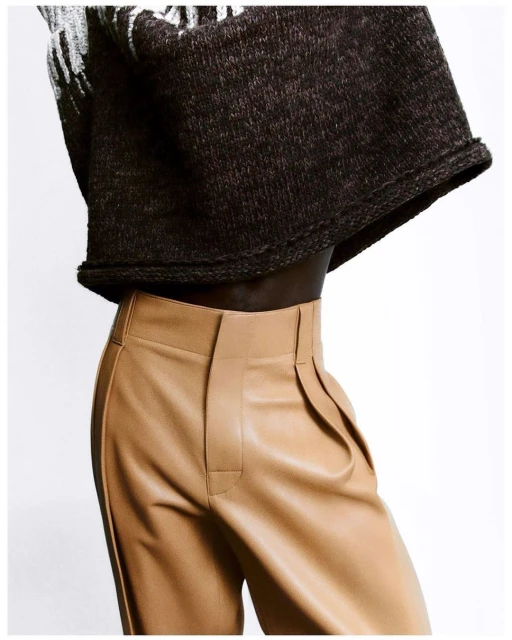 Close-up fashion shot of a torso wearing a black patterned sweater and high-waisted tan trousers against a white background.