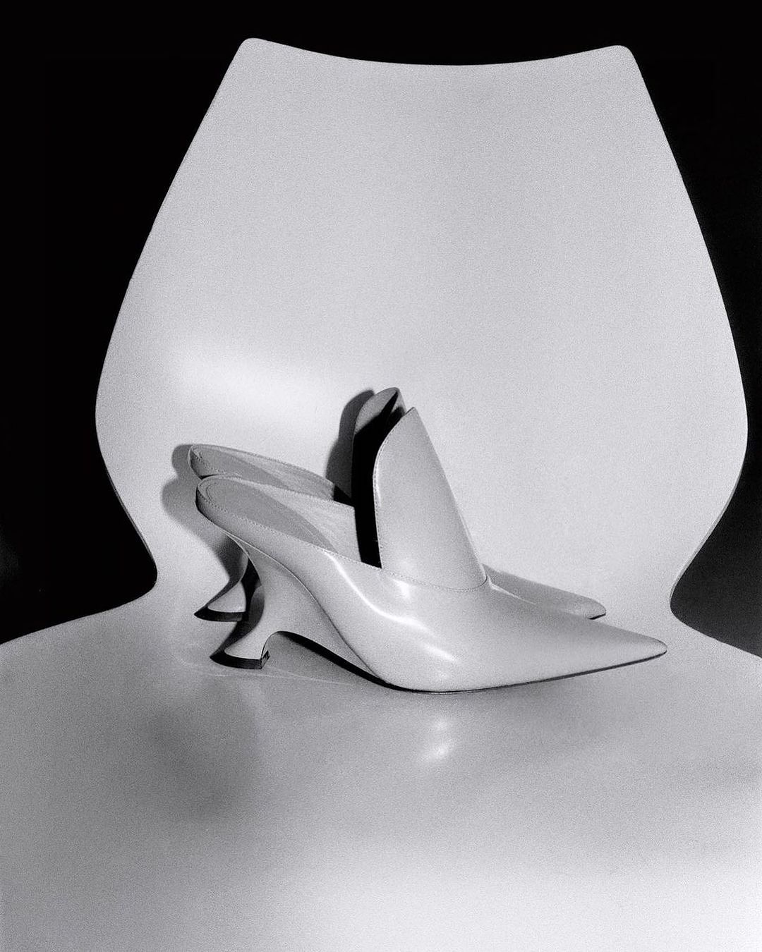 Ivory-colored heeled mules arranged on a glossy white chair against a dark backdrop, showcasing minimalist shoe design.
