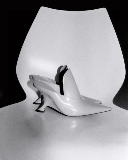 Ivory-colored heeled mules arranged on a glossy white chair against a dark backdrop, showcasing minimalist shoe design.