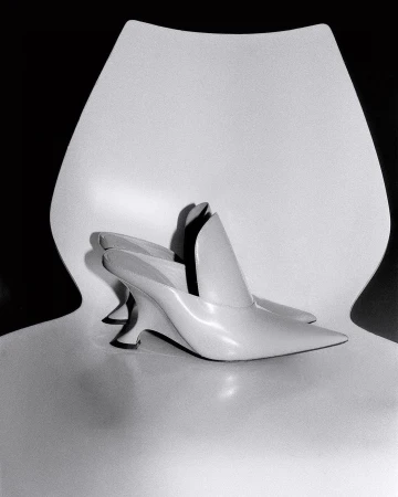 Ivory-colored heeled mules arranged on a glossy white chair against a dark backdrop, showcasing minimalist shoe design.