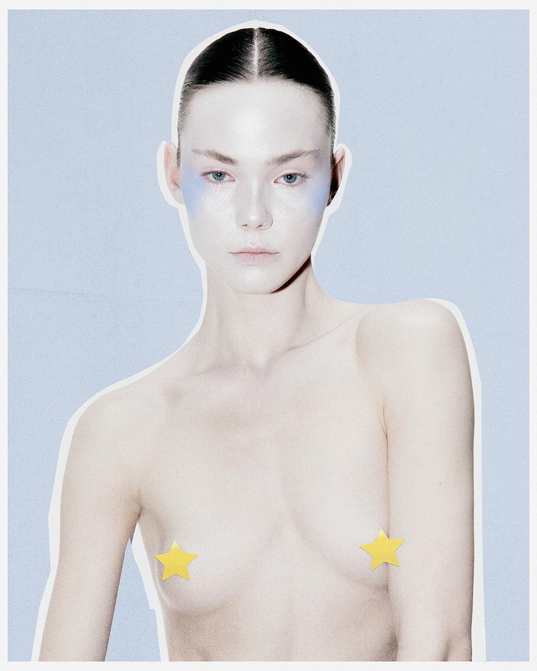 Conceptual beauty shot of a topless adult woman with blue face makeup and yellow star nipple pasties against a light blue background.