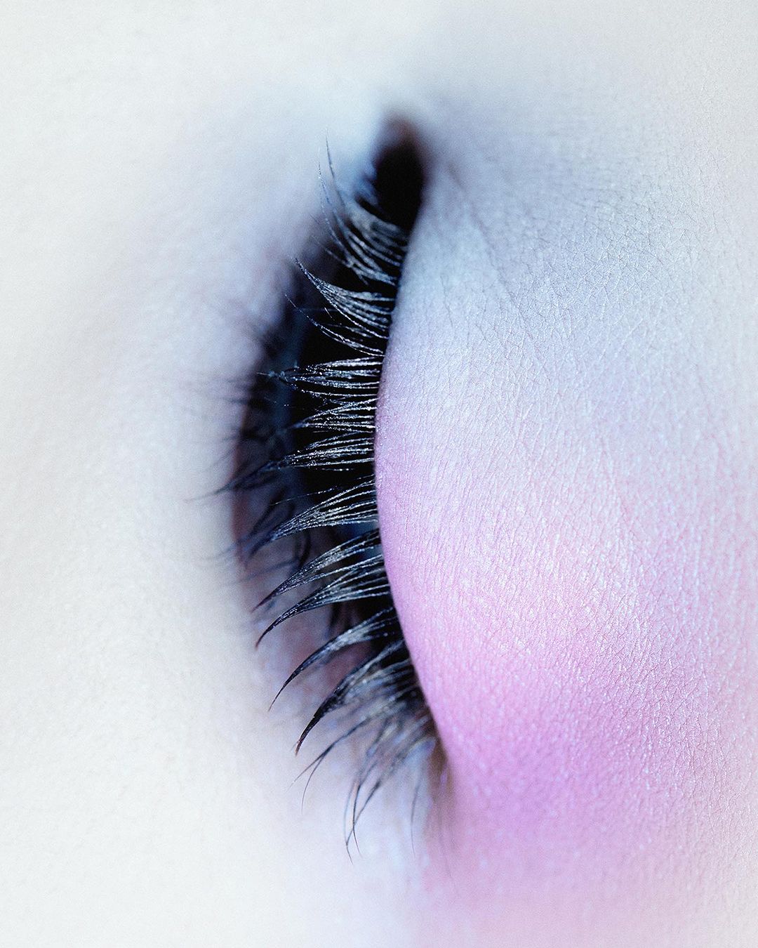 Macro shot of a closed eye with long, dark eyelashes and light skin with soft pink eyeshadow, showcasing beauty and makeup artistry.