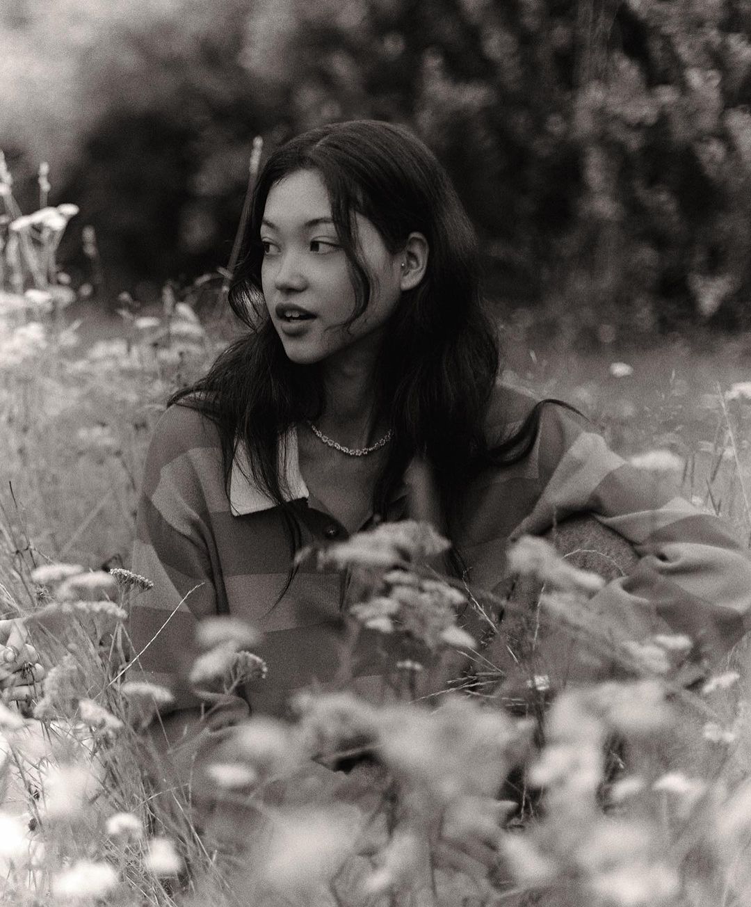 Monochrome portrait of a young woman with dark hair, wearing a striped shirt, sitting in a field of flowers, looking off to the side.