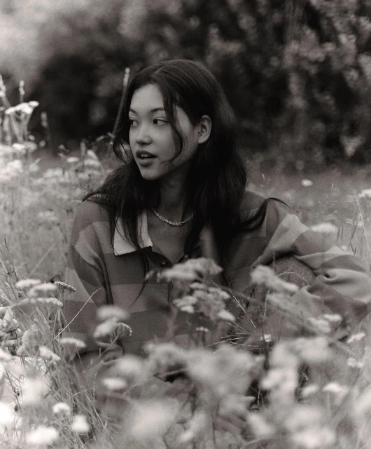 Monochrome portrait of a young woman with dark hair, wearing a striped shirt, sitting in a field of flowers, looking off to the side.