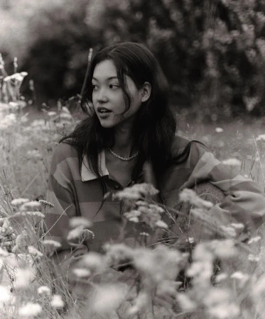Monochrome portrait of a young woman with dark hair, wearing a striped shirt, sitting in a field of flowers, looking off to the side.