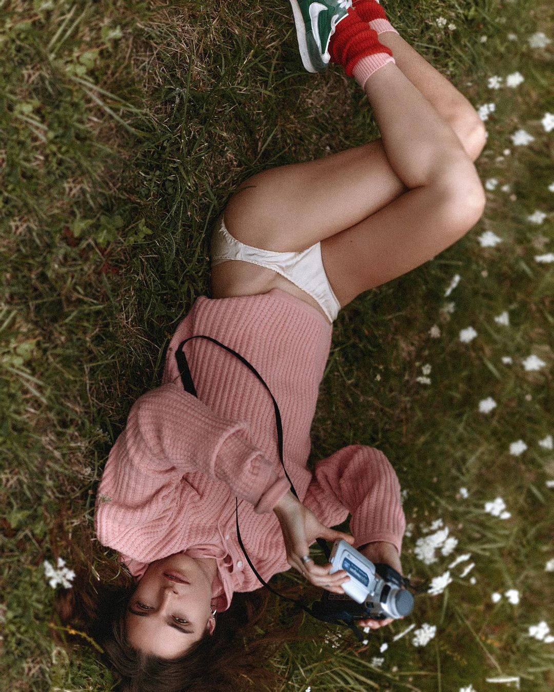 Young woman lying on grass, wearing a pink sweater, socks, and underwear, holding a retro camcorder in an outdoor setting.