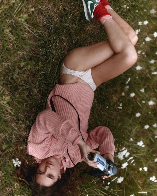 Young woman lying on grass, wearing a pink sweater, socks, and underwear, holding a retro camcorder in an outdoor setting.