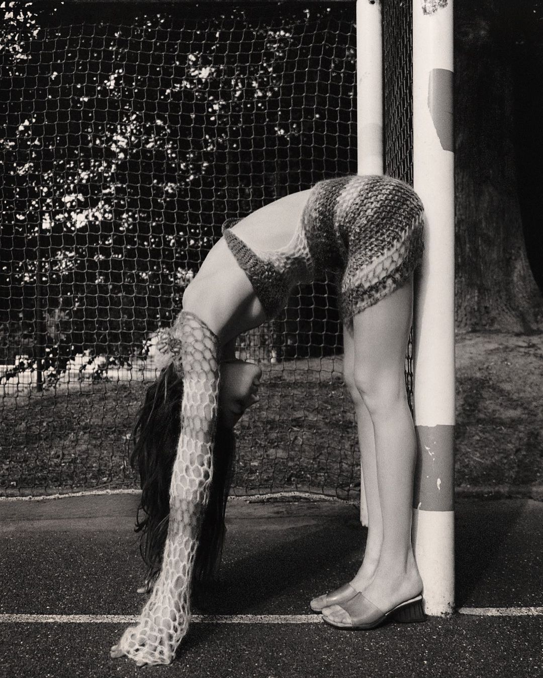 Woman in a knit outfit bends backward against a goalpost on an outdoor sports field in a black-and-white shot with a net background.