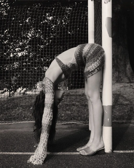 Woman in a knit outfit bends backward against a goalpost on an outdoor sports field in a black-and-white shot with a net background.