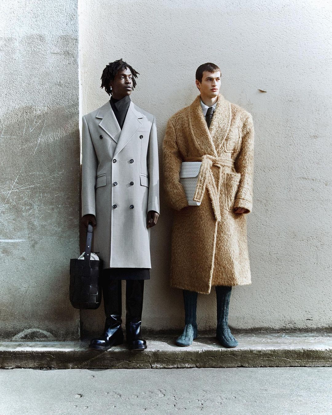 Two male fashion models in coats pose in an urban setting, one in grey double-breasted coat and the other in a tan robe coat.