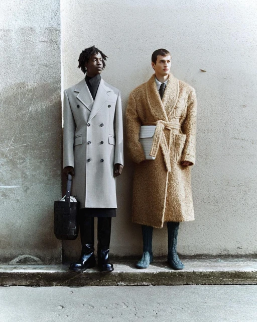 Two male fashion models in coats pose in an urban setting, one in grey double-breasted coat and the other in a tan robe coat.