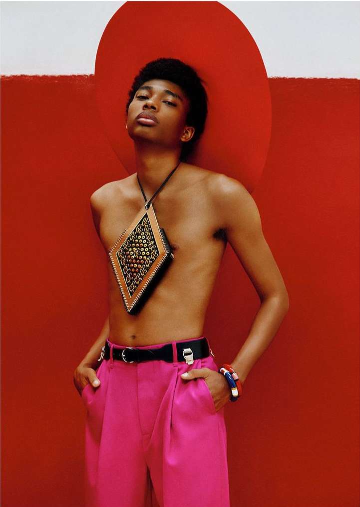 Model in magenta pants poses against a red and white background with a diamond-shaped purse; fashion portrait in photo studio setting.