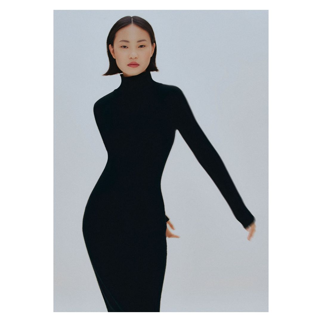 A slender Asian woman posing in a sleek black turtleneck dress against a bright white backdrop in a minimalist studio fashion shot.