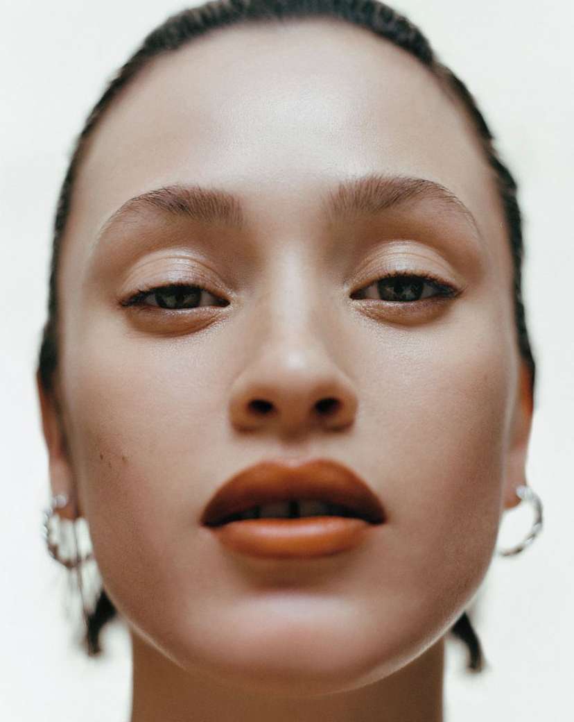 Close-up portrait of an adult female model with caramel lips, natural make-up, and silver earrings, set against a soft, pale background.