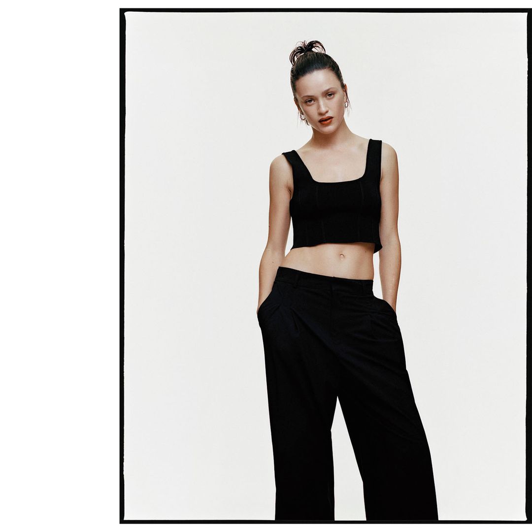 Portrait of a woman wearing a black cropped top and dark trousers with hands in pockets against a white studio backdrop.