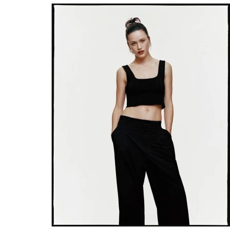 Portrait of a woman wearing a black cropped top and dark trousers with hands in pockets against a white studio backdrop.