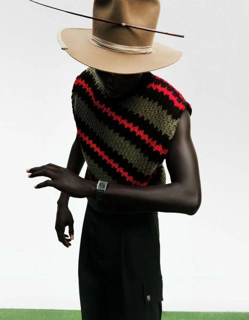 Stylish studio shot of a Black male fashion model wearing a knit vest and tan hat with a thin bow-like structure, posing elegantly against a neutral backdrop.
