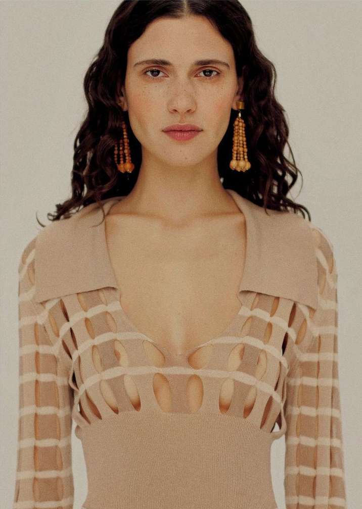 A woman with dark curly hair and dangling earrings poses in a studio, wearing a beige knit top with a cut-out detail.