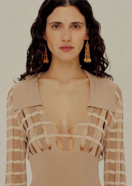 A woman with dark curly hair and dangling earrings poses in a studio, wearing a beige knit top with a cut-out detail.