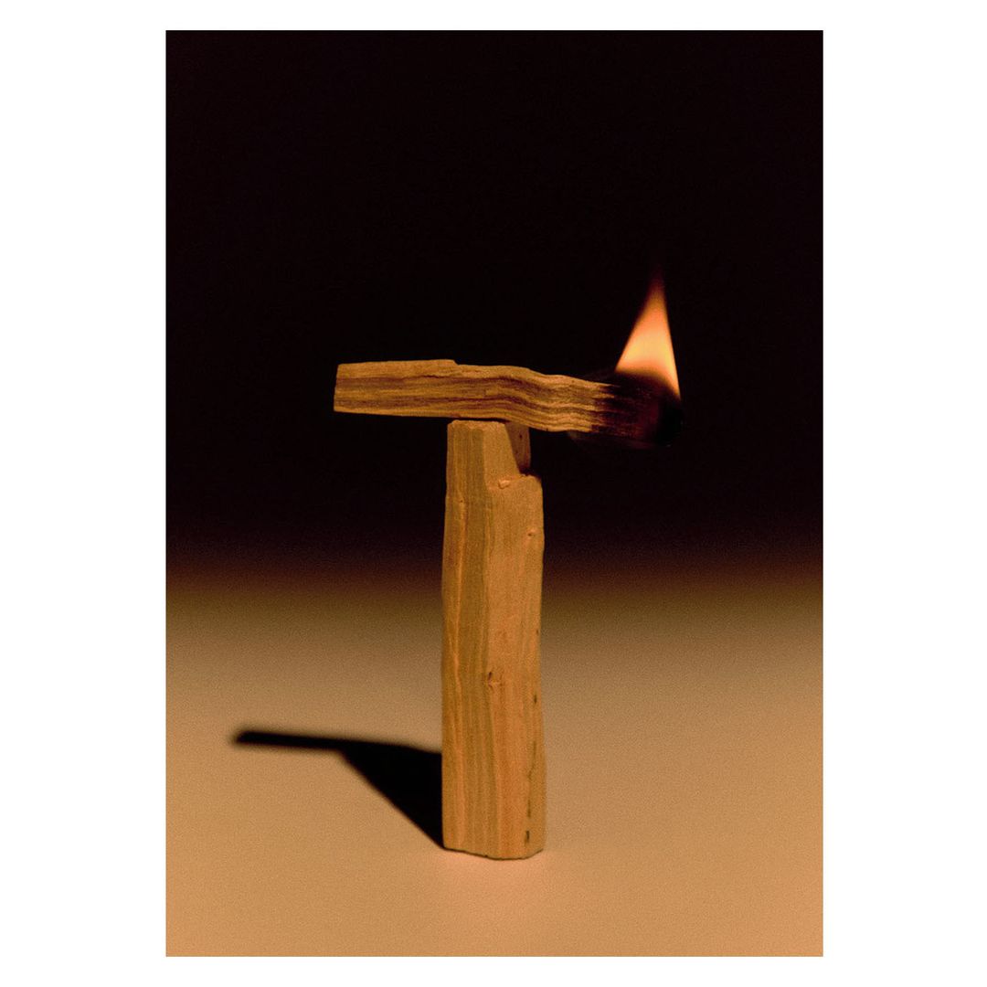 Lit palo santo wood resting vertically on a table; a small flame burns on one end. The background is dark and out of focus.