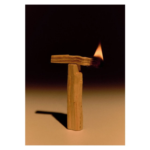 Lit palo santo wood resting vertically on a table; a small flame burns on one end. The background is dark and out of focus.
