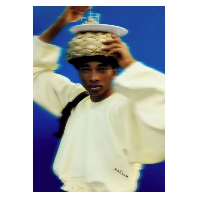 Person balancing decorative basket, glasses, and plate on head in a studio setting against a blue background, wearing a cream sweatshirt.