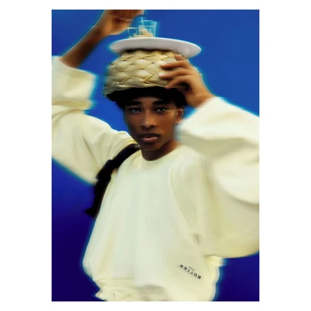 Person balancing decorative basket, glasses, and plate on head in a studio setting against a blue background, wearing a cream sweatshirt.