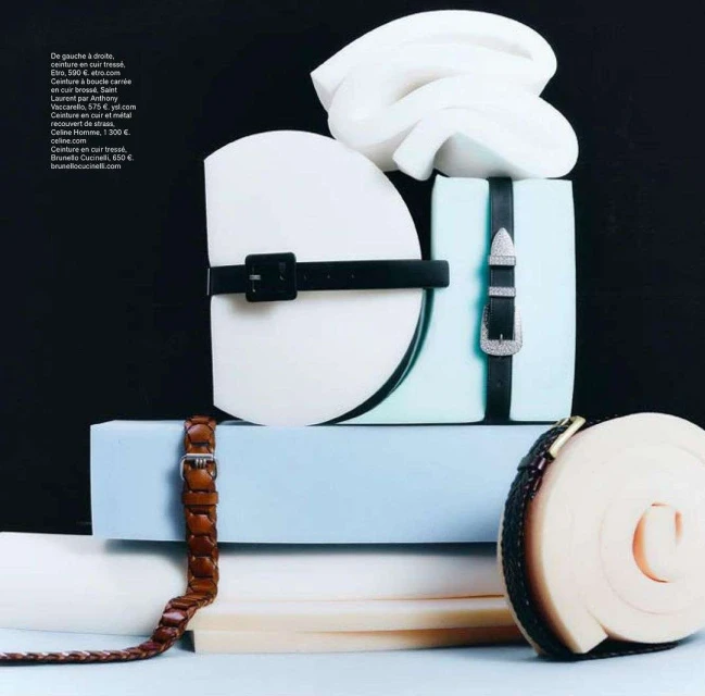 Still life of assorted belts with buckles, artfully arranged on stacked white and light blue blocks against a black backdrop.