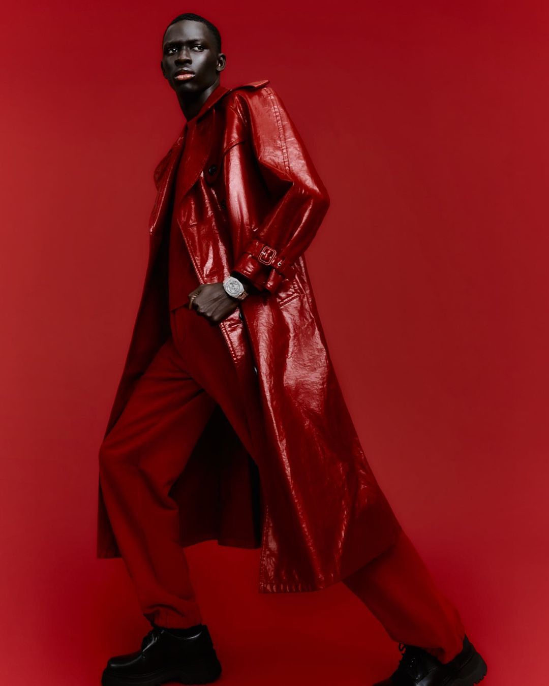 Fashionable man in a glossy red trench coat and trousers against a matching backdrop, showcasing modern style and texture.