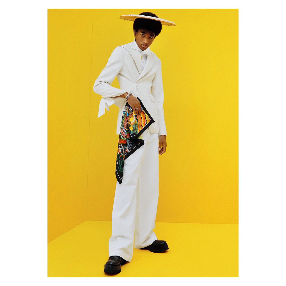 A person in a white suit and hat stands in front of a bright yellow background, holding a colorful patterned scarf, in a studio shot.