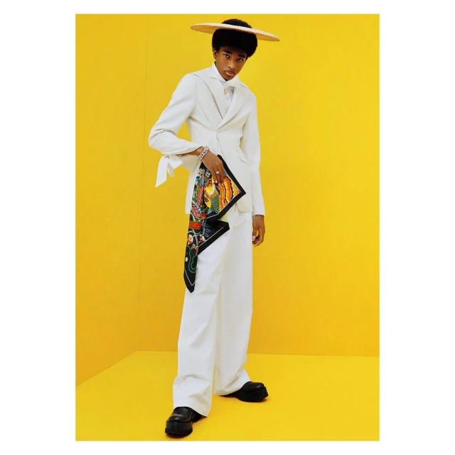 A person in a white suit and hat stands in front of a bright yellow background, holding a colorful patterned scarf, in a studio shot.