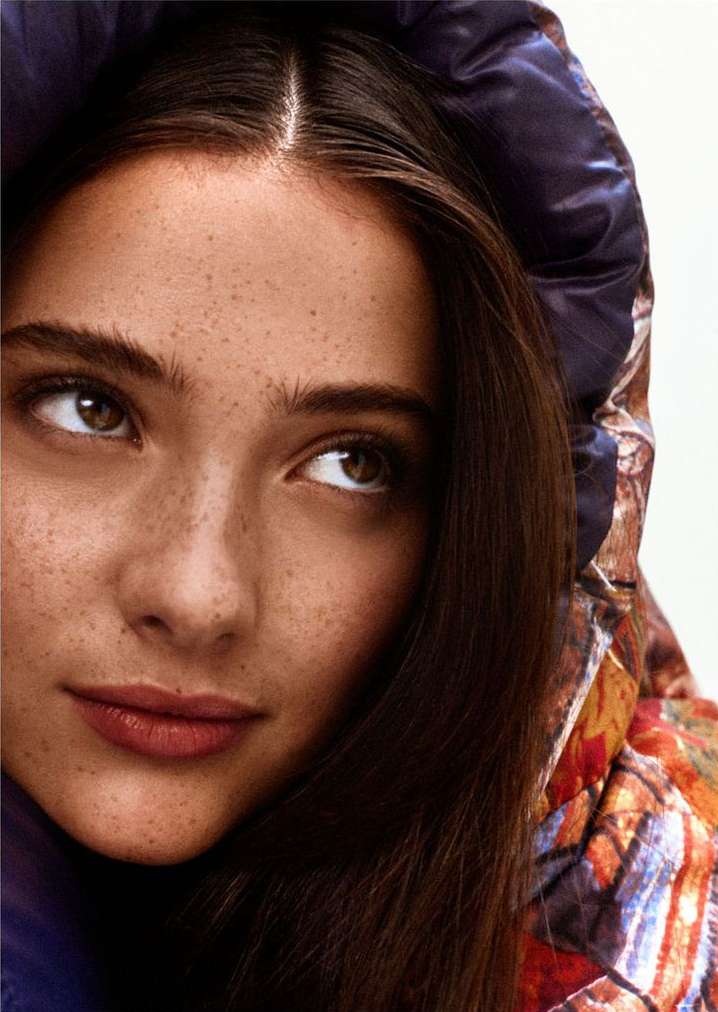 Close-up portrait of a young adult woman with auburn hair and freckled skin wearing a hooded jacket, looking off to the side.