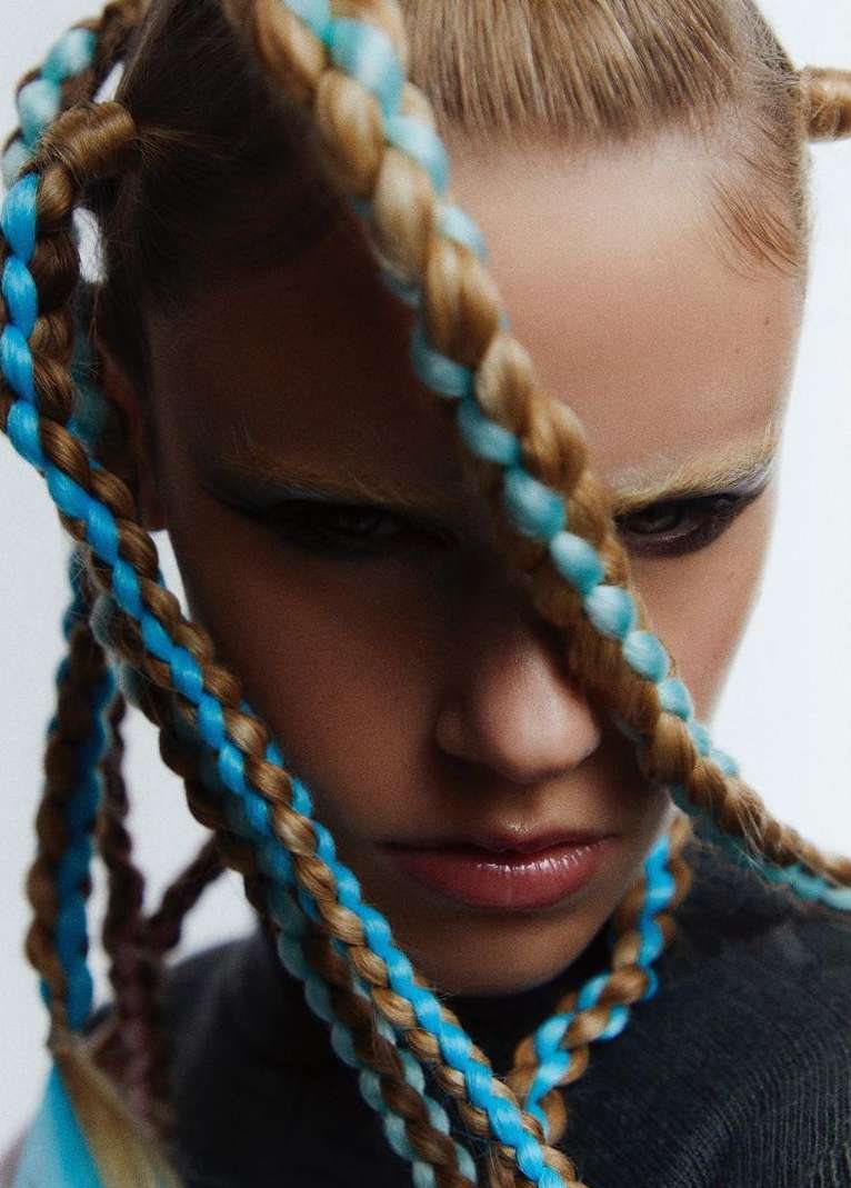Close-up studio shot of an adult female model with blonde hair styled in braided pigtails with blue extensions, wearing a gray top.