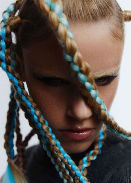 Close-up studio shot of an adult female model with blonde hair styled in braided pigtails with blue extensions, wearing a gray top.