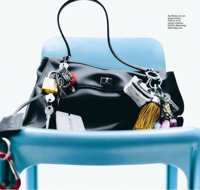 Black Balenciaga handbag sitting on a light blue chair with multiple keychains attached in a studio setting with a white backdrop.