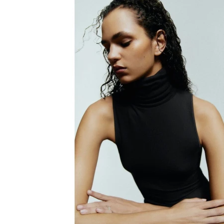 A young adult woman with curly hair poses in a sleeveless turtleneck top against a neutral background in a close up shot.