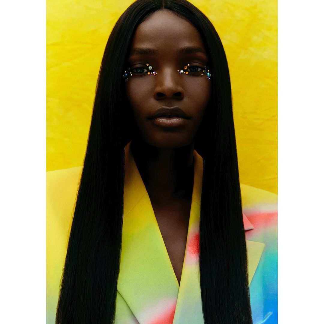 Striking beauty shot of a Black woman with jewel makeup, dark hair and a yellow-to-pastel-color blazer against a bright yellow backdrop.