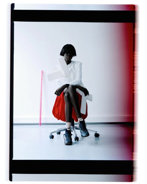 Dark-skinned adult female fashion model in studio, seated on wheeled office chair wearing designer sneakers, white top, and red skirt.