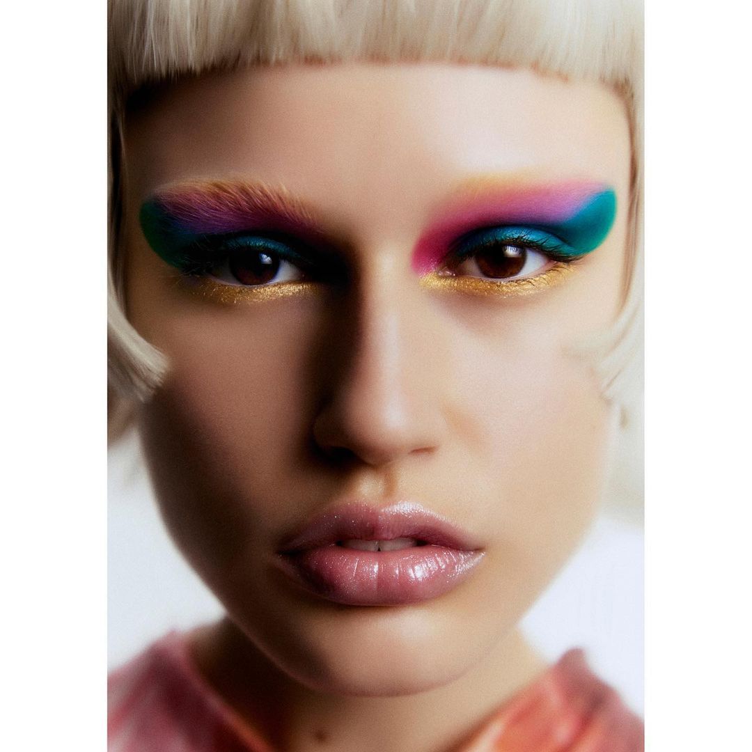 Close-up portrait of a woman with colorful eyeshadow, pink lipstick, and blonde bangs against a plain white background.