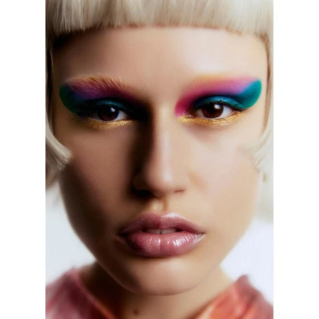 Close-up portrait of a woman with colorful eyeshadow, pink lipstick, and blonde bangs against a plain white background.