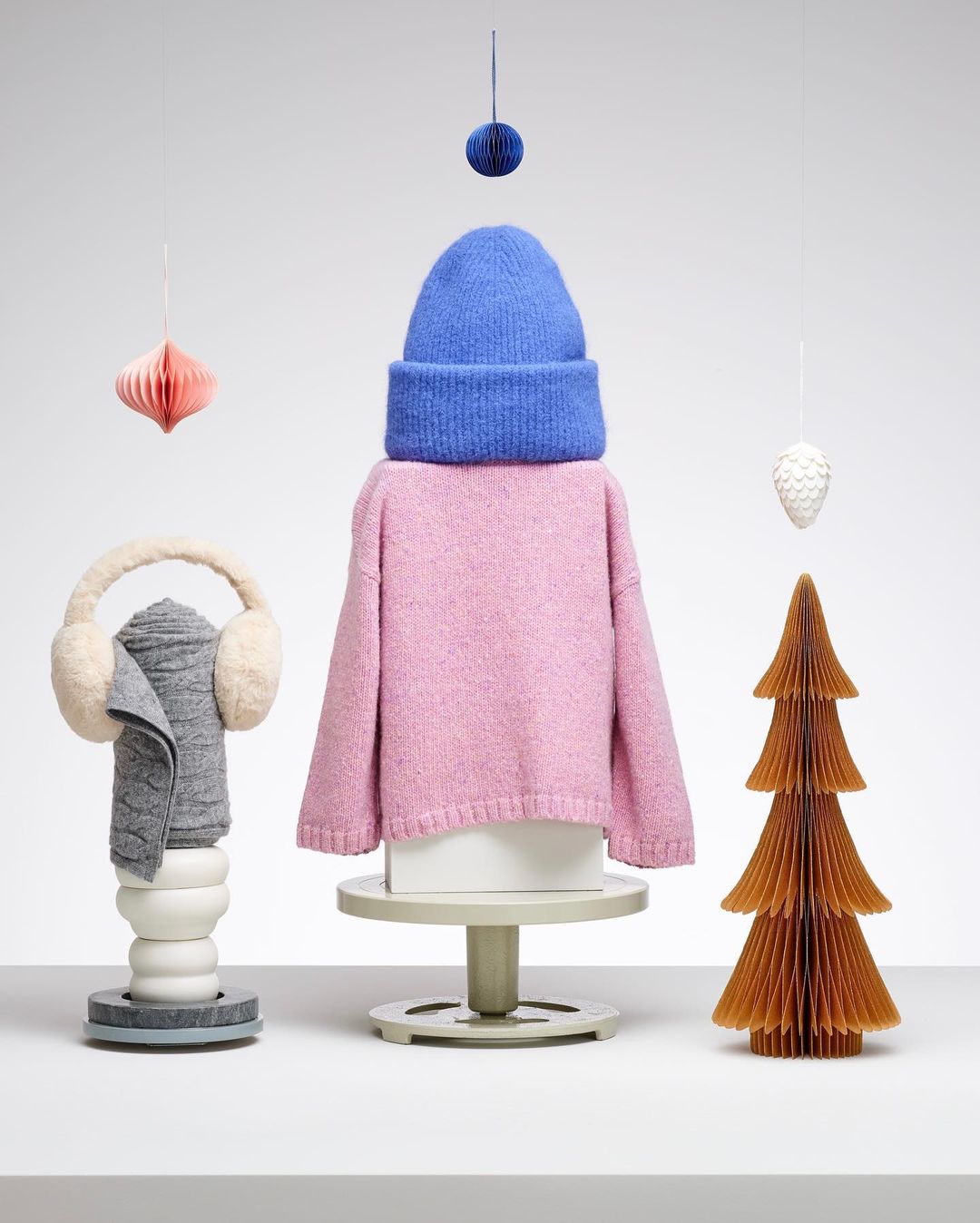 Stylized product display features a pink sweater, blue beanie, earmuffs, and paper ornaments arranged on pedestals against a white background.