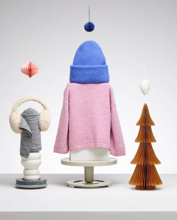 Stylized product display features a pink sweater, blue beanie, earmuffs, and paper ornaments arranged on pedestals against a white background.