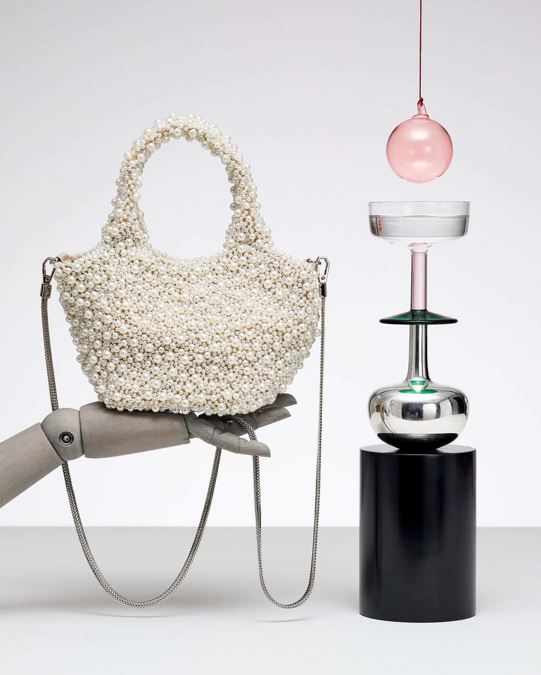 Pearl-encrusted handbag held by mannequin hand next to artistic glassware on a cylindrical pedestal in a minimalist studio setting.